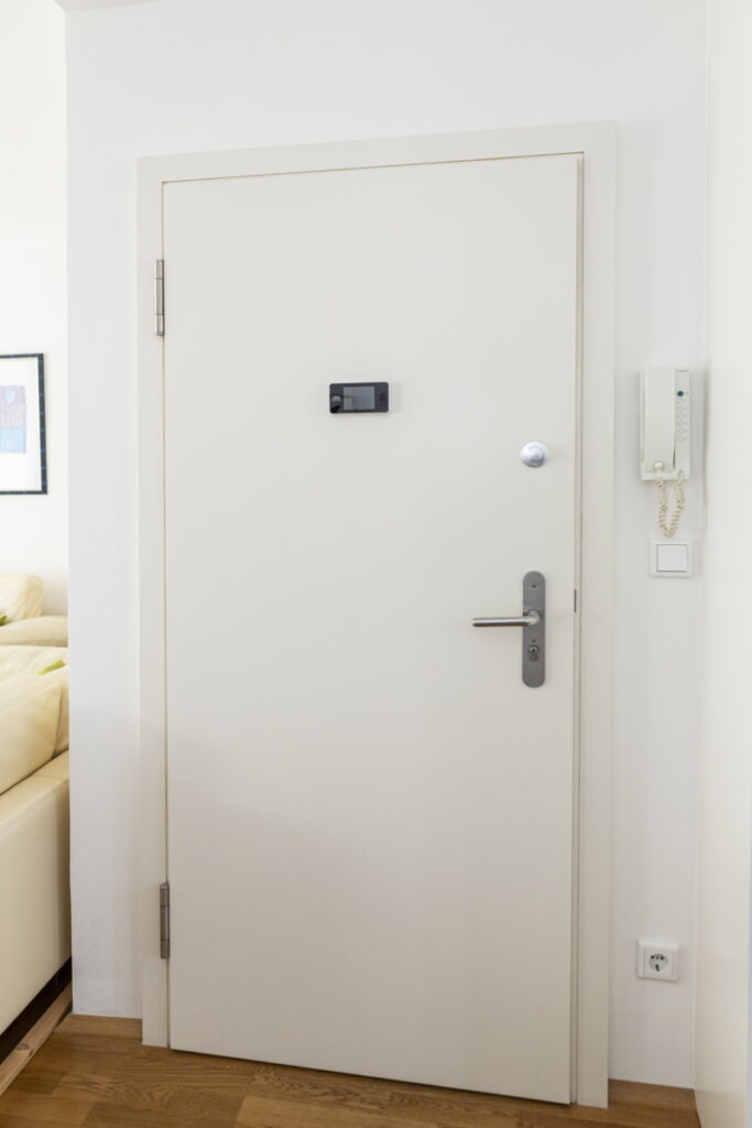 Sichere Wohnungstür mit moderner Technik Weiße Wohnungseingangstür mit Türspion, modernem Schloss und Gegensprechanlage an der Wand daneben.