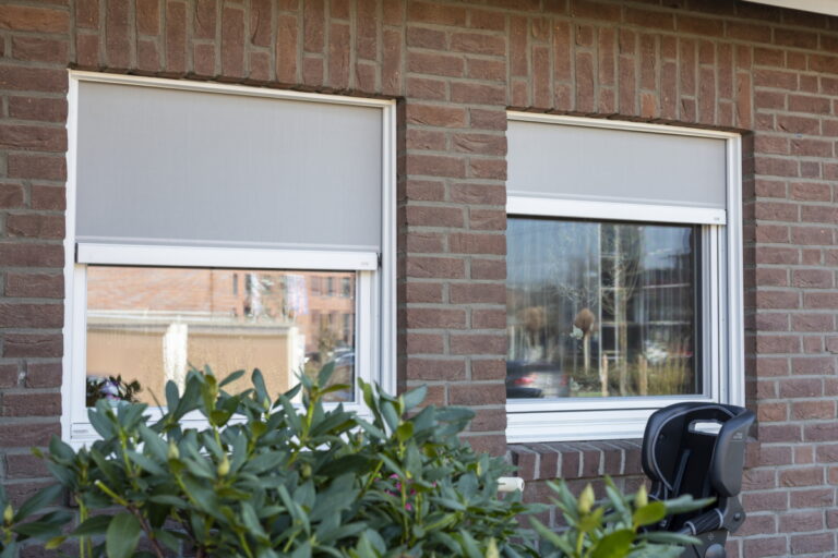 ROMA 5 Zwei Fenster mit Insektenschutz-Spannrahmen in einer roten Klinkerfassade, davor grüne Sträucher und ein Kinderautositz.