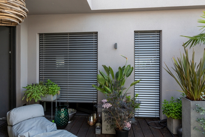 ROMA 3 Kleine Terrasse mit modernen Außenraffstores, verschiedenen Pflanzen und einem Lounge-Sessel.