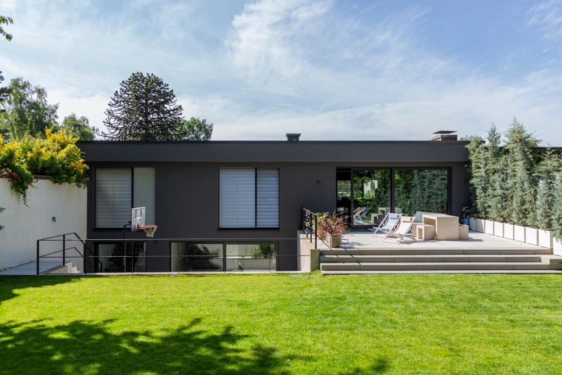 KOWA 4 Modernes Flachdachhaus mit dunkler Fassade, großer Terrasse, Liegestühlen und gepflegtem Garten mit Rasenfläche.