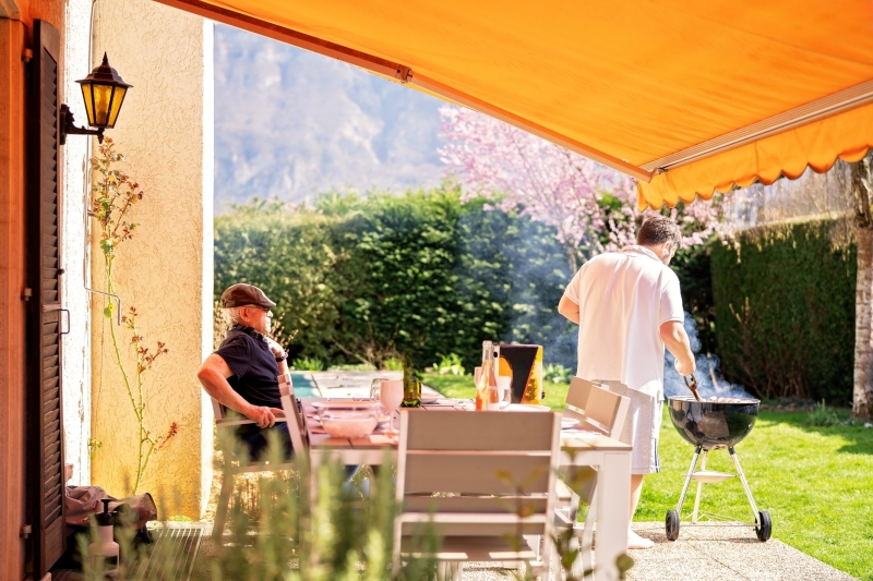 markilux 1 Zwei Männer genießen einen sonnigen Tag im Garten unter einer orangefarbenen Markise, einer sitzt am Tisch, der andere grillt.