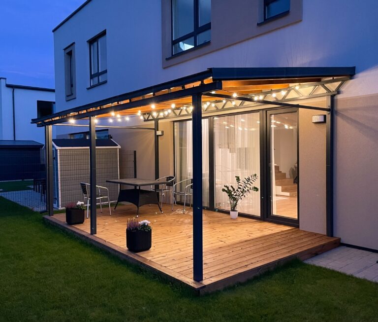 Moderne Terrassenüberdachung mit Beleuchtung – stilvolles Wohnen im Freien Moderne Terrasse mit Holzbelag, Glasüberdachung und stimmungsvoller Beleuchtung an einem Einfamilienhaus bei Abenddämmerung.