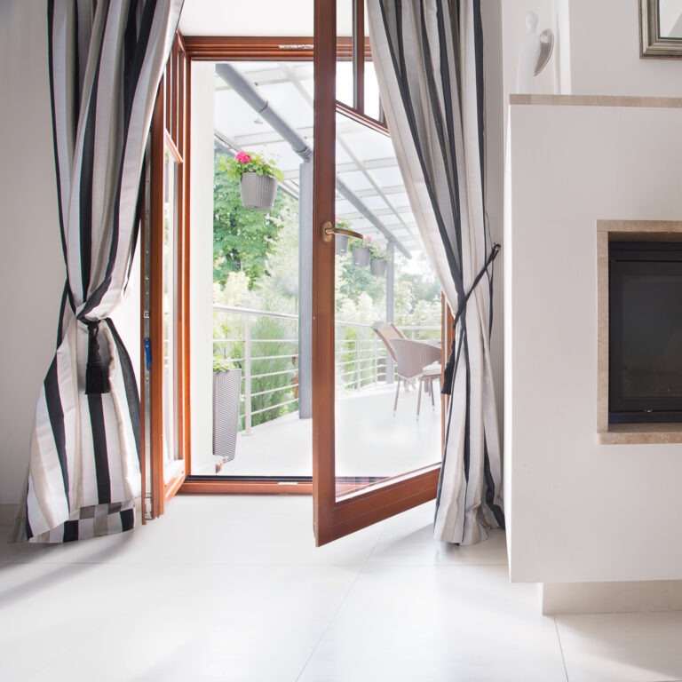 Heller Wohnraum mit Zugang zur Terrasse – Wohnkomfort mit Aussicht Geöffnete Holztür führt vom Wohnzimmer auf einen hellen Balkon mit Vorhängen und hängenden Pflanzen.