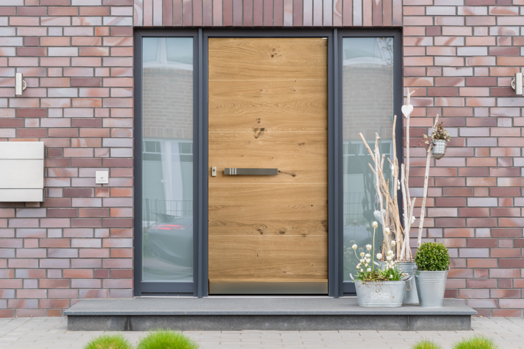 GRAUTHOFF 4 Moderner Hauseingang mit heller Holzhaustür, seitlichen Glasflächen und dekorativen Pflanzgefäßen vor einer roten Klinkerfassade.