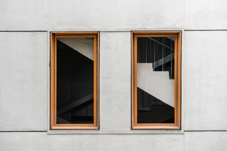 Moderne Architektur – klare Linien und Betonfassade mit Holzfenstern Zwei rechteckige Fenster mit Holzrahmen in einer grauen Betonfassade, durch die ein Treppenhaus sichtbar ist.