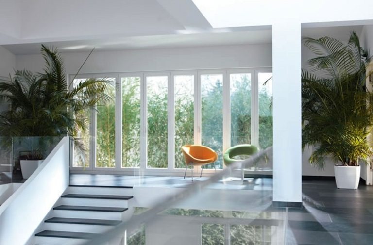 Weru 3 Helles modernes Treppenhaus mit großen weißen Fenstern, Palmen in Pflanzkübeln und zwei farbigen Sesseln.