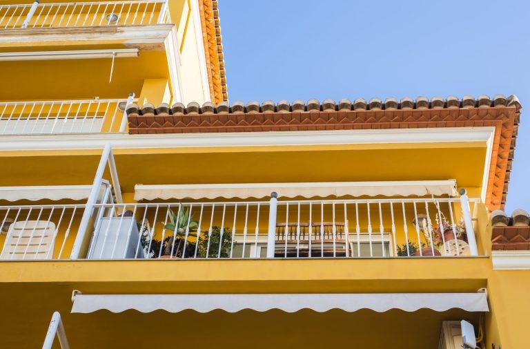 Mediterranes Wohnhaus mit Markisen und sonnigem Balkon Gelbes Wohnhaus mit Balkonen, weißen Geländern und ausgefahrener Markise unter blauem Himmel.